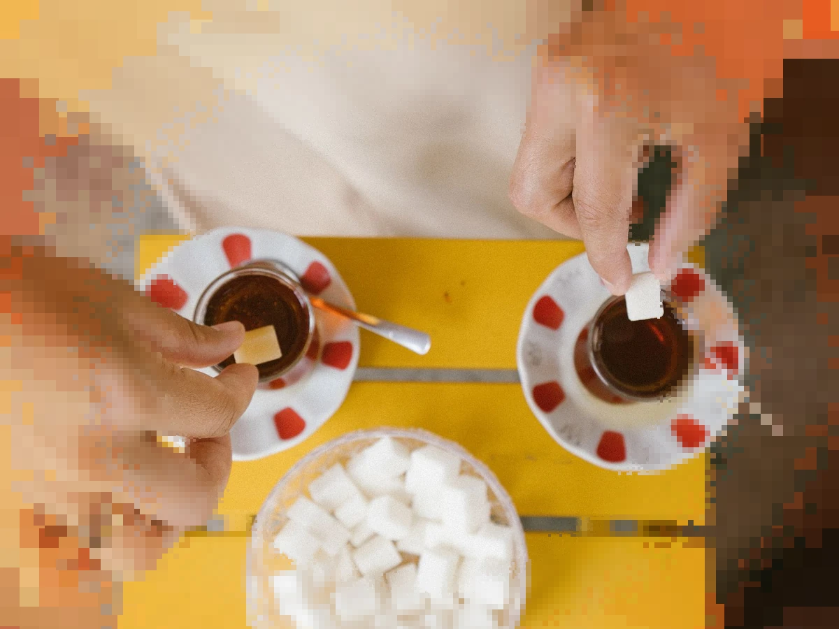 Two hands drop sugar cubes into small glasses of tea on red patterned saucers, with a bowl of sugar cubes on a yellow table, the edges breaking into pixels