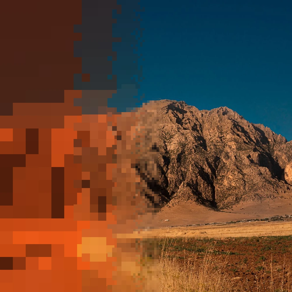 A bare, rocky mountain in evening light above dry grass, the left side breaking into ember pixels