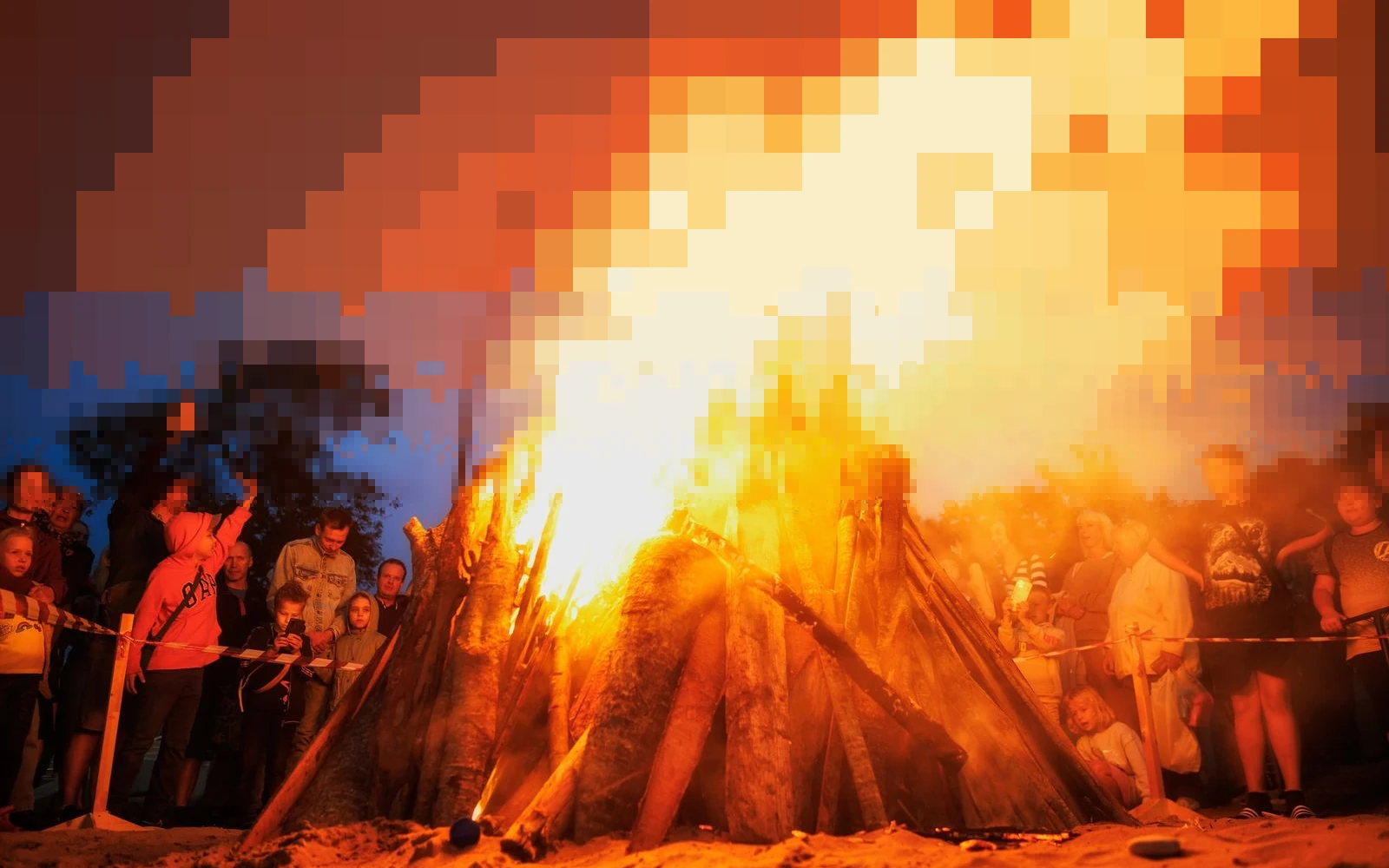 A tall bonfire blazing at dusk with a crowd watching from behind a rope, the flames breaking into ember pixels toward the top