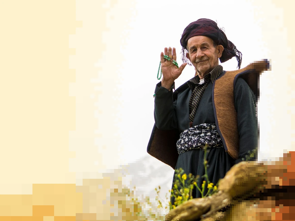 An older Kurdish man in a dark turban, a felt waistcoat and a patterned sash raises his hand in greeting with prayer beads, sharp around him and pixelated toward the edges